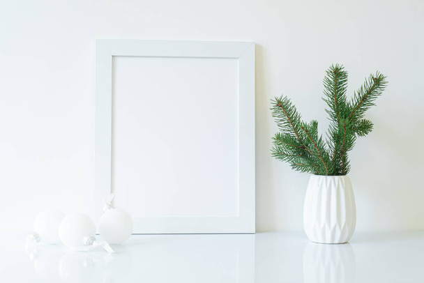 Mock up white frame with fir branch in vase and white christmas decorations on shelf. Empty portrait frame against a white wall - Photo, image
