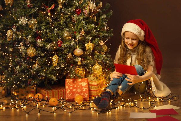 Christmas Child in Santa Hat holding Greeting Card Letter. Little Girl Sitting Next to Gift Stacks and Xmas Tree. Winter Holiday Night Home Interior Decoration - Foto, imagen