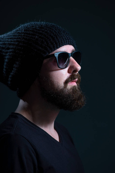 authentic portrait of a bearded millennial in sunglasses and a black knitted hat against a dark background. - Fotoğraf, Görsel