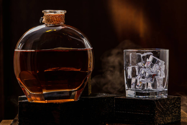 Black varnished wooden table, with a whiskey bottle and glass with ice, black background with smoke, yellow light - Photo, image
