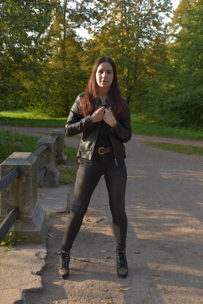 Brunette girl in a black leather jacket and black jeans in an autumn park - Фото, изображение