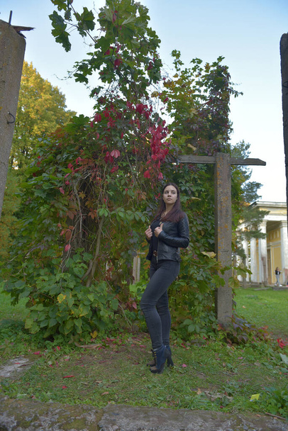 Brunette girl in a black leather jacket and black jeans in an autumn park - Фото, изображение