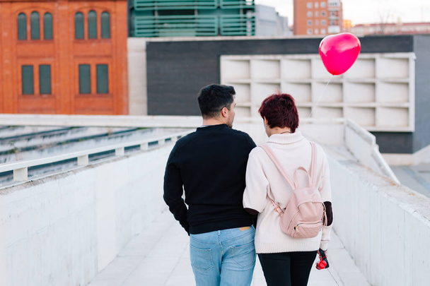 Δύο άνθρωποι, ένα νεαρό ζευγάρι, περπατούν προς τα πίσω μέσα στην πόλη, αγκαλιάζοντας ένα μπαλόνι σε σχήμα κόκκινης καρδιάς την ημέρα του Αγίου Βαλεντίνου. Έννοια των εραστών και της πρώτης αγάπης. - Φωτογραφία, εικόνα