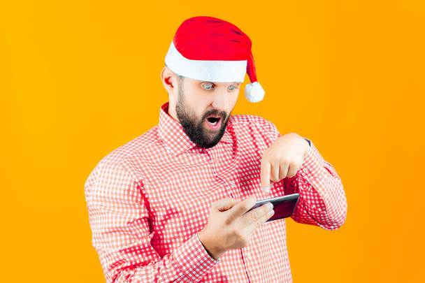 ein Mann mit Neujahrsmütze vom Weihnachtsmann blickt überrascht auf das Telefon, öffnet den Mund und zeigt mit dem Finger darauf. - Foto, Bild