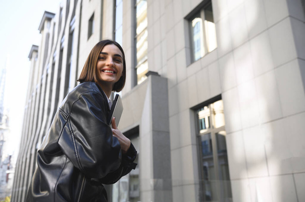 Successful self-confident attractive woman, business person, freelancer, entrepreneur, copywriter holding laptop in her hands and cutely smiles looking at camera against corporate buildings background - Photo, Image