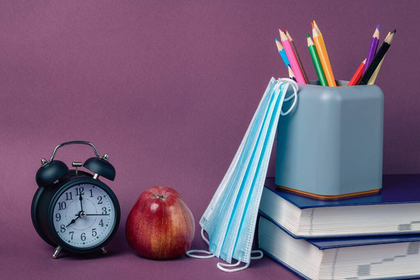 Черный будильник, красное яблоко, медицинские маски и школьные принадлежности на фиолетовом фоне. В школу, коронавирусные ограничения - Фото, изображение