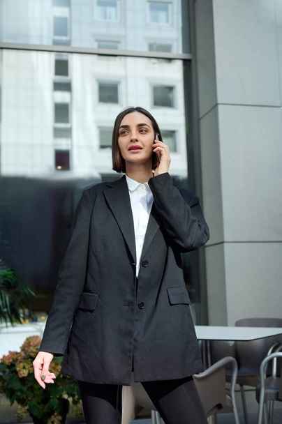 Attractive freelance woman with stylish short haircut, successful business person dressed in fashionable casual clothes smiles talking on mobile phone on modern corporate high-rise building background - Φωτογραφία, εικόνα
