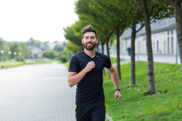 Young Caucasian bearded happy man runner jogging down the street on urban background of city park. Morning jog. Active healthy lifestyle fitness. Sport workout outdoor. Athlete training run exercise - Foto, Imagen