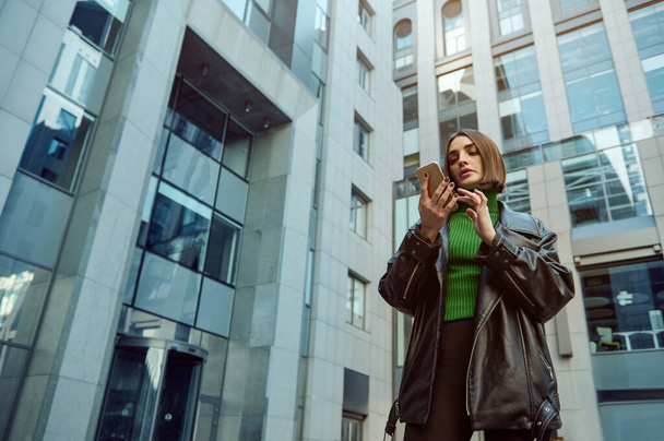 Beautiful self-confident European young woman, freelancer, entrepreneur, copywriter using mobile phone on modern office corporate high-rise buildings background. Business and communications concept. - Фото, зображення