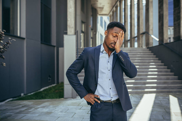 exausto jovem empresário afro-americano trabalhador de escritório ficar ao ar livre. homem preto sobrecarregado estressado com uma cabeça massagens de dor de cabeça. Deprimido Empregado cansado sofrendo dor trabalho doente fora - Foto, Imagem