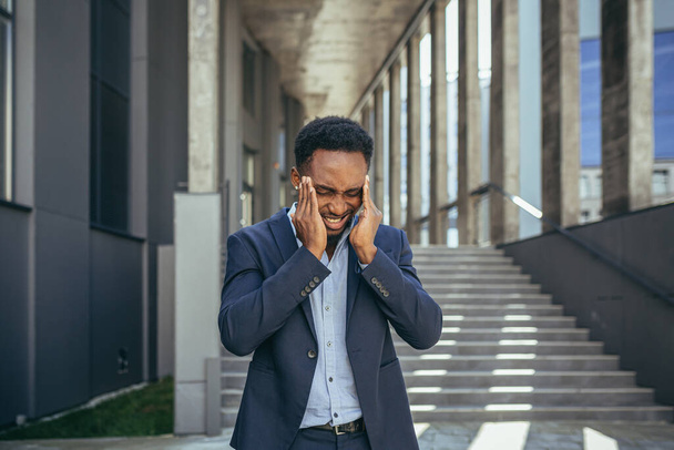 exhausted young African American business man office worker stand outdoors. stressed overworked black male with a headache massages head. Depressed Tired employe suffering pain sick work outside - Fotografie, Obrázek