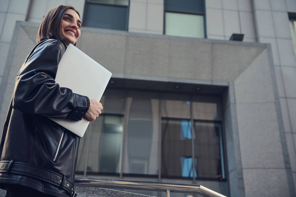 Με αυτοπεποίθηση κίνητρα επιτυχημένη νεαρή γυναίκα atractive freelancer περπάτημα με ένα φορητό υπολογιστή στα χέρια, χαμόγελα κοιτάζοντας πέρα από τη σύγχρονη εταιρική επιχειρηματικό κέντρο φόντο - Φωτογραφία, εικόνα