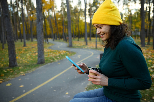 Šťastná usměvavá dospělá žena v teplých pestrobarevných šatech sedící na lavičce v parku a držící odnášející papírový kelímek, používající mobilní telefon a vychutnávající si podzimní počasí odpočívající v podzimním dubovém háji - Fotografie, Obrázek