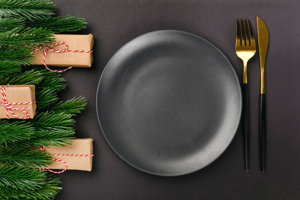 vista dall'alto di coltelleria nera e forchetta vicino piatto vuoto nero su tavolo nero con rami di albero di Natale e decorazioni da scatole regalo - Foto, immagini