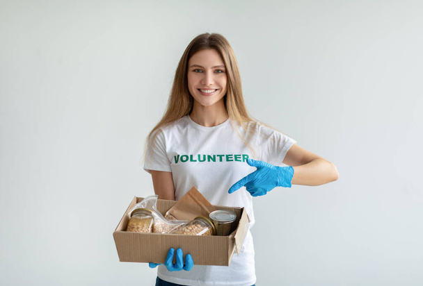Felice volontaria femminile in possesso e indicando scatola con donazioni, sorridendo alla macchina fotografica, in piedi su sfondo chiaro - Foto, immagini