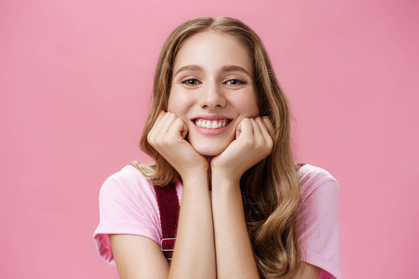 Cute feminine and tender nice girl withtattoo and wavy hair leaning head on hands as if listening amusing story gazing with broad smile and admiration gaze being dreamy over pink background - Фото, изображение