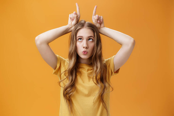 GIrl going crazy from boredom making freaky funny faces fooling around squinting holding index fingers on head like horns sticking out tongue and folding lips aping over orange background - Photo, Image