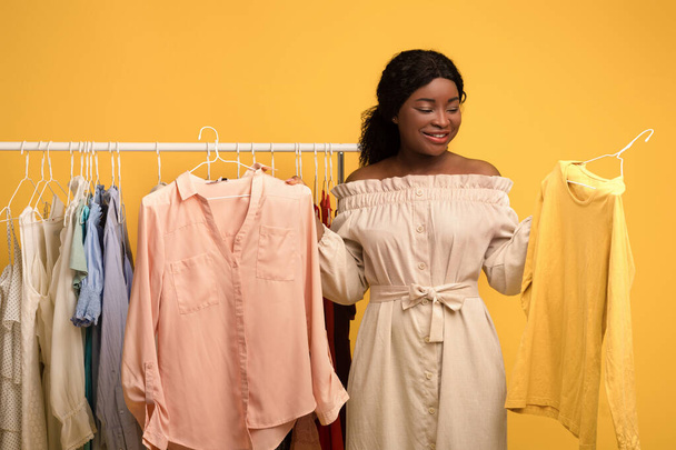 Černá žena stojící u stojanu na šaty se stylovým oblečením, vybírá si mezi dvěma oblečky, nakupuje módní šatník - Fotografie, Obrázek
