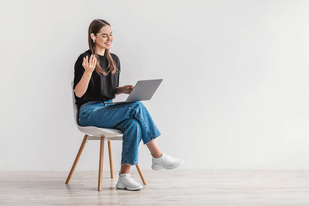 Χαρούμενη νεαρή γυναίκα σε ακουστικά που χρησιμοποιεί φορητό υπολογιστή, έχει online συνάντηση, κάθεται στην καρέκλα ενάντια στο λευκό τοίχο, κενό χώρο - Φωτογραφία, εικόνα