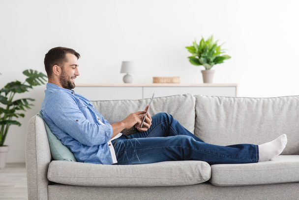 Alegre europeo adulto chico en casual relajarse en el sofá, navegar en Internet y charlar en tiempo libre - Foto, Imagen