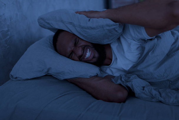 Stresli siyah adam, kulaklarını kapatıyor. Geceleri yatakta yatıyor. - Fotoğraf, Görsel