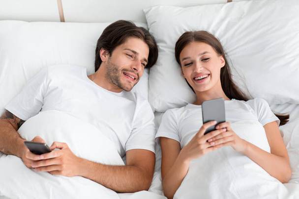Veselý pohledný tisíciletý Evropan muž se dívá na manželku smartphone v posteli v ložnici interiér - Fotografie, Obrázek