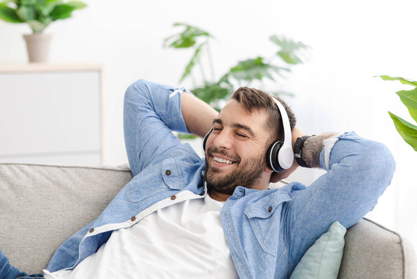 Satisfied european adult guy in casual and headphones resting on couch, listening music at free time - Foto, immagini