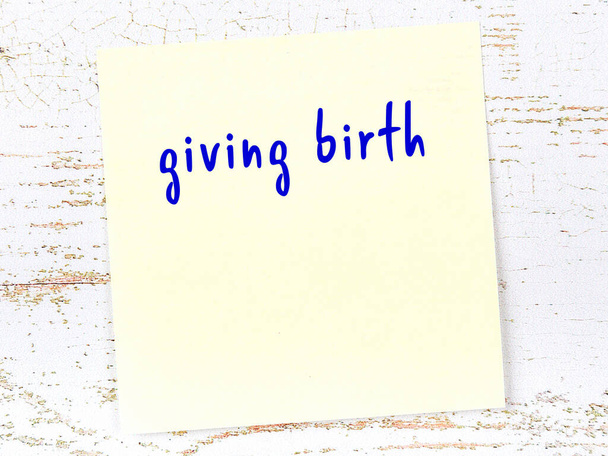 Yellow sticky note on wooden wall with handwritten inscription giving birth - Photo, Image