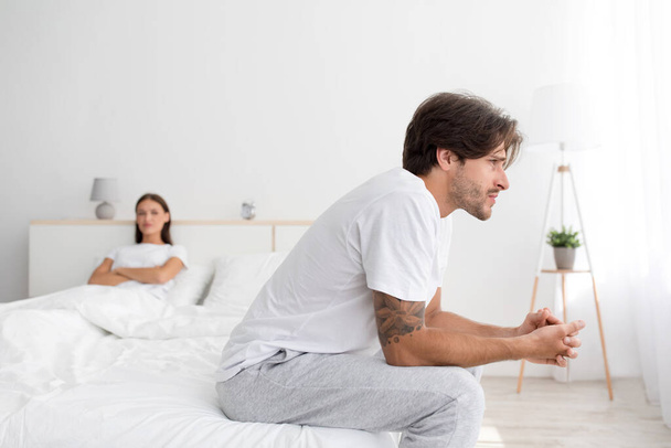 Smutny obrażony młody biały mąż siedzi na łóżku, ignorując żonę z skrzyżowanymi ramionami w sypialni - Zdjęcie, obraz