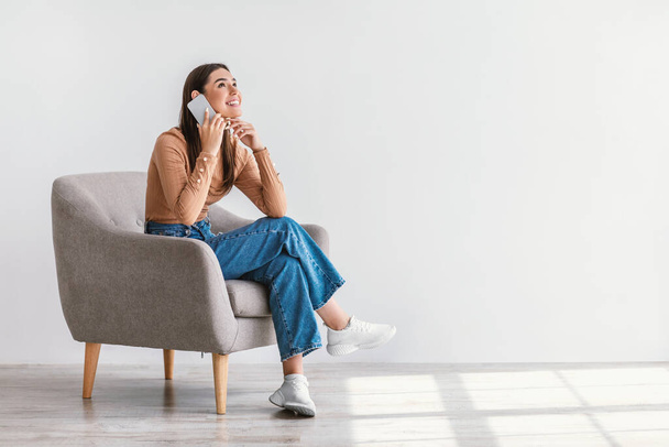 Neşeli genç bir kadın cep telefonuyla konuşuyor, koltuğunda oturuyor, beyaz duvara karşı hoş bir telefon konuşması yapıyor. - Fotoğraf, Görsel