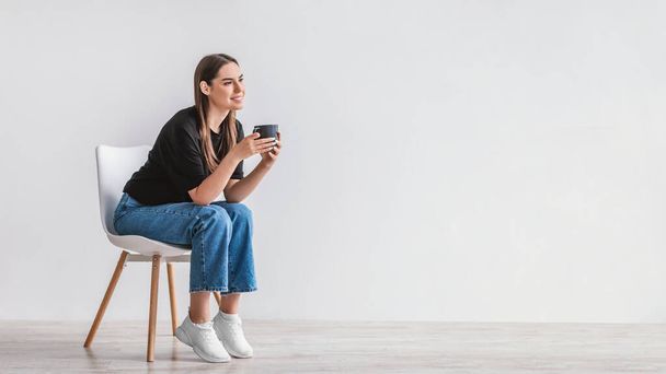 Coffee break. Dreamy young woman sitting on chair, enjoying cup of hot beverage against white wall, copy space - Фото, изображение