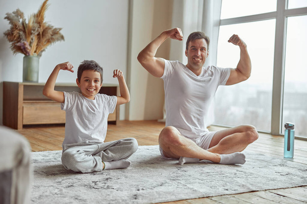 Joyful boy demonstrating muscles with dad at home - 写真・画像