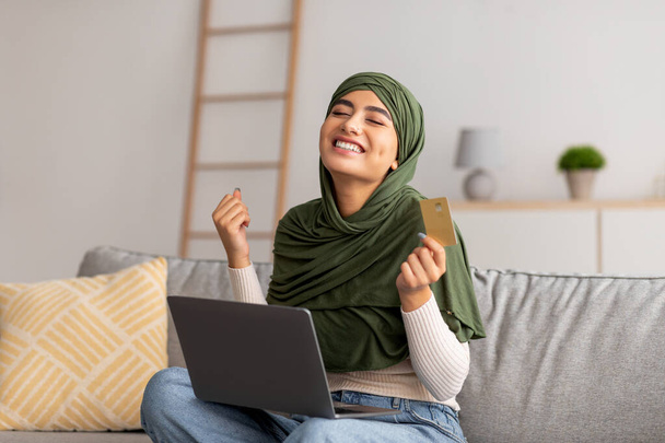 ラップトップを使用して、クレジットカードを保持しているヒジャーブの感情的な若いアラブ人女性は、はいジェスチャーを作る、自宅でオンラインショッピング - 写真・画像