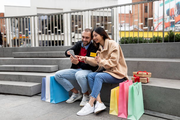 Mladý pár sedí na schodech s chytrým telefonem a kreditní kartou, nakupuje na webu, obklopen nákupními taškami a dárky - Fotografie, Obrázek