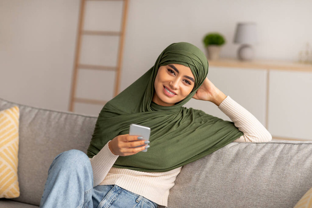 Mujer árabe sonriente en hijab sentado en el sofá, el uso de teléfonos inteligentes, navegar por la web, charlando en línea, viendo la película en casa - Foto, imagen