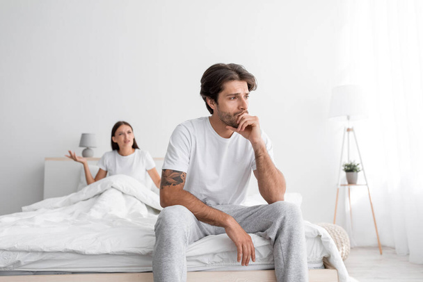 Depressieve duizendjarige blanke man met stoppels zit op bed, boze vrouw schreeuwt en zweert in slaapkamer interieur - Foto, afbeelding