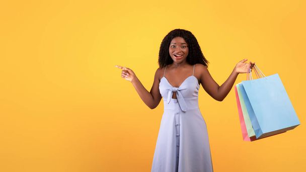Fröhliche junge schwarze Frau mit bunten Einkaufstaschen, die zur Seite zeigen und großen Verkauf oder Promo auf orangefarbenem Hintergrund anbieten - Foto, Bild
