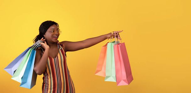 Fröhliche schwarze Dame im Kleid hält Einkaufstüten in der Hand und zeigt zur Seite auf den leeren Raum auf orangefarbenem Studiohintergrund - Foto, Bild