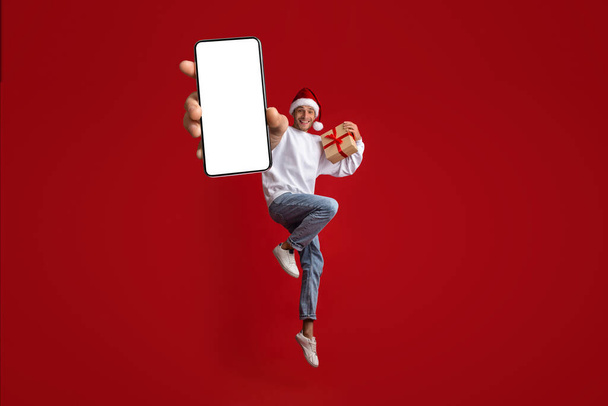 Vtipný chlap s vánoční dárek v ruce skákání s velkým prázdným Smartphone - Fotografie, Obrázek