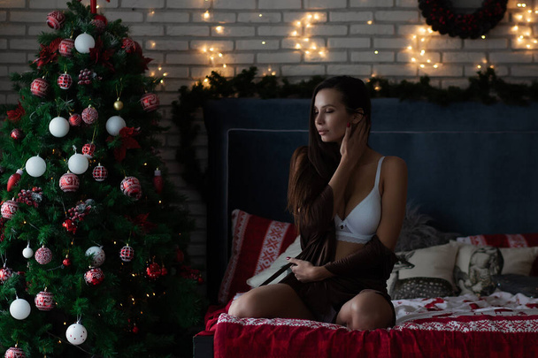 Senhora jovem auto-assegurada em robe de seda marrom e sutiã branco sentado em cama confortável em quarto acolhedor decorado com guirlandas brilhantes e grinalda durante as férias de Natal - Foto, Imagem