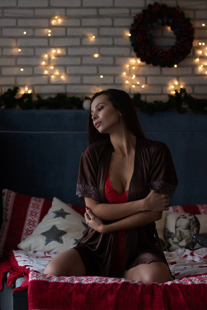 Αυτοδιαβεβαίωση νεαρή κοπέλα σε καφέ μεταξωτή ρόμπα και κόκκινο bodysuit κάθεται σε άνετο κρεβάτι στο άνετο δωμάτιο διακοσμημένα με λαμπερό γιρλάντες και στεφάνι κατά τη διάρκεια των διακοπών των Χριστουγέννων - Φωτογραφία, εικόνα