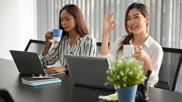Šťastná žena zaměstnanec gestikulace ok ruční podepsat, zatímco její kolegové pití kávy a práce na notebooku - Fotografie, Obrázek