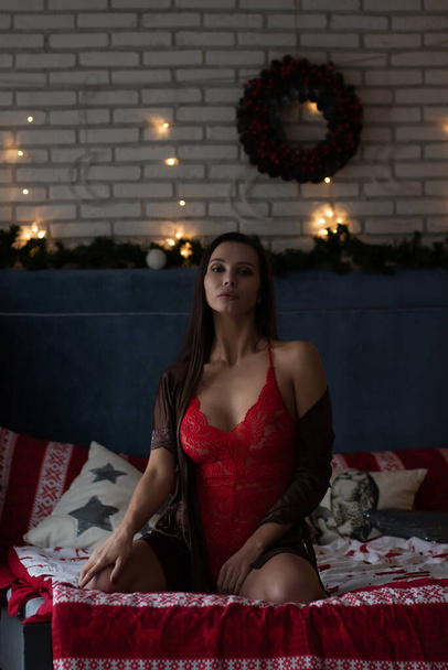 Autosicuro giovane signora in abito di seta marrone e body rosso seduto su un comodo letto in camera accogliente decorato con ghirlande incandescenti e ghirlanda durante le vacanze di Natale - Foto, immagini