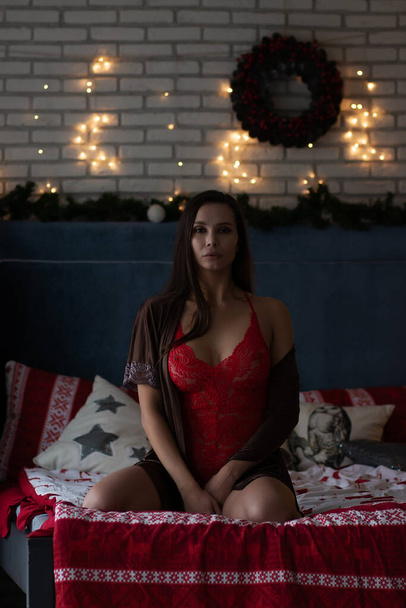 Jovencita segura de sí misma en bata de seda marrón y traje de cuerpo rojo sentada en una cómoda cama en una acogedora habitación decorada con guirnaldas brillantes y corona de flores durante las vacaciones de Navidad - Foto, Imagen