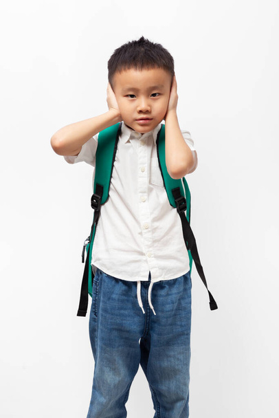 Kind mit Schulrucksack, der die Ohren schließt, trägt weißes Hemd auf weißem Hintergrund - Foto, Bild