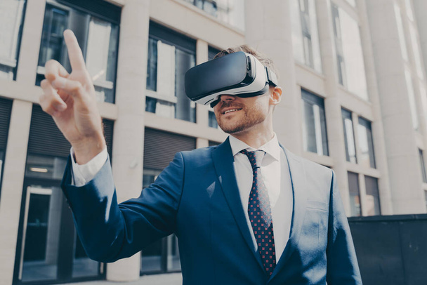 Vrolijke zakenman probeert iets aan te raken in virtual reality, staan in de buurt van kantoorgebouw - Foto, afbeelding