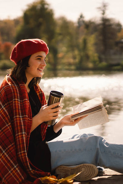 Nastolatka w czerwonym berecie czytająca książkę o drewnianym pontonie. Jesienny sezon. - Zdjęcie, obraz