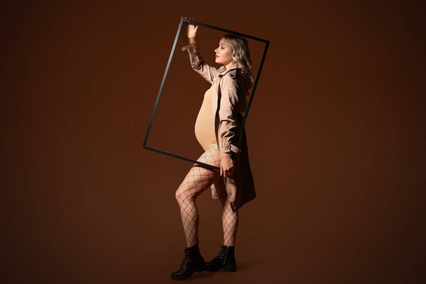 Stylish young pregnant woman with frame on brown background - Φωτογραφία, εικόνα