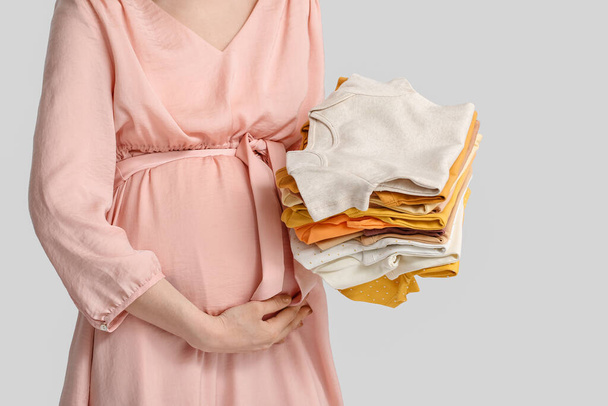 Young pregnant woman with stack of baby clothes on light background - Photo, image