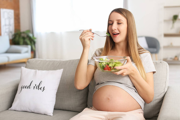 Young pregnant woman eating vegetable salad on sofa at home - 写真・画像